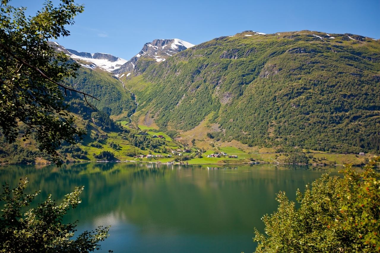 Kandal, Gloppen -Green mountains with snow patches rise above a calm lake, with a small village on the shore under blue sky..