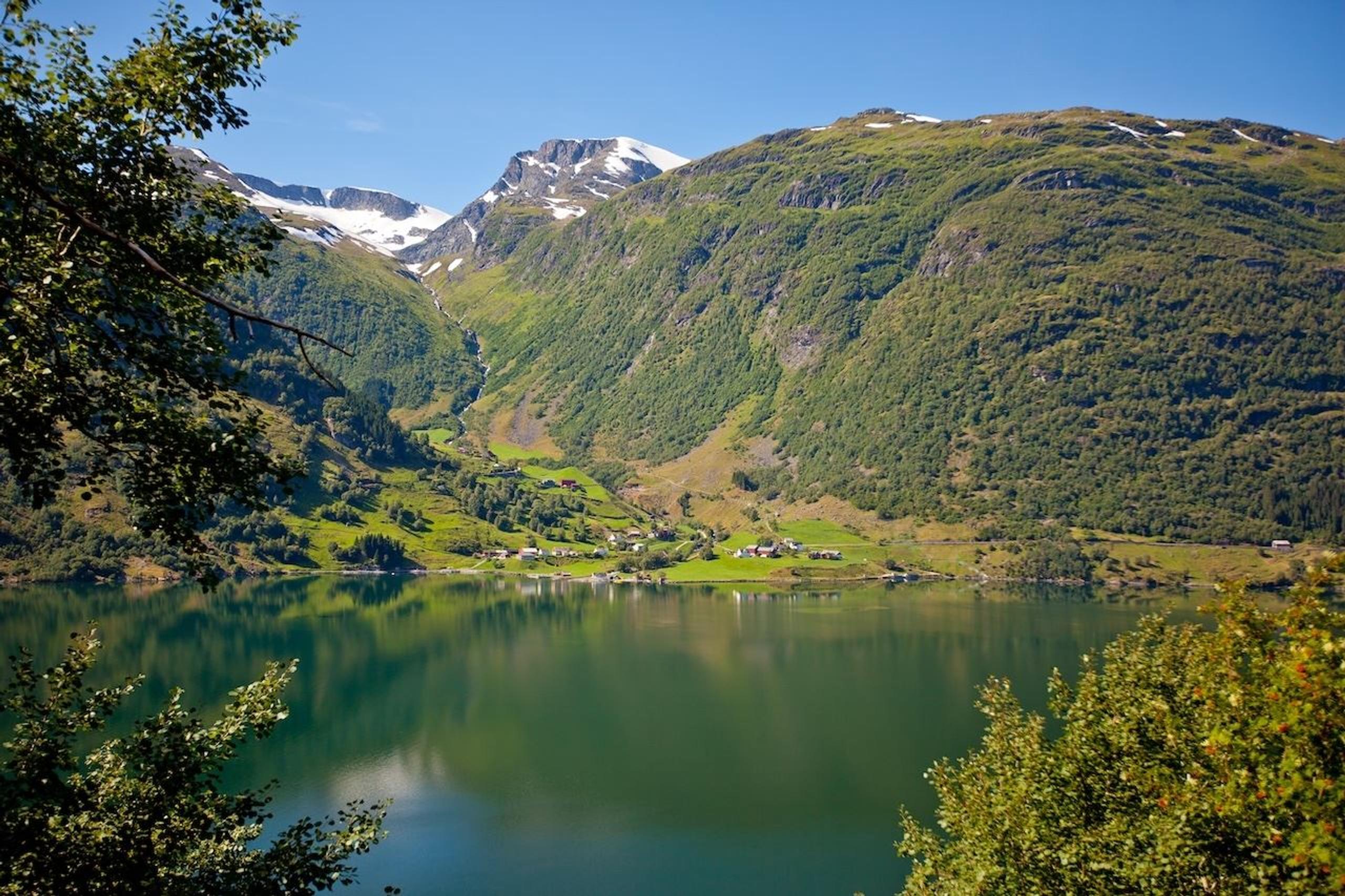 Kandal, Gloppen -Green mountains with snow patches rise above a calm lake, with a small village on the shore under blue sky..