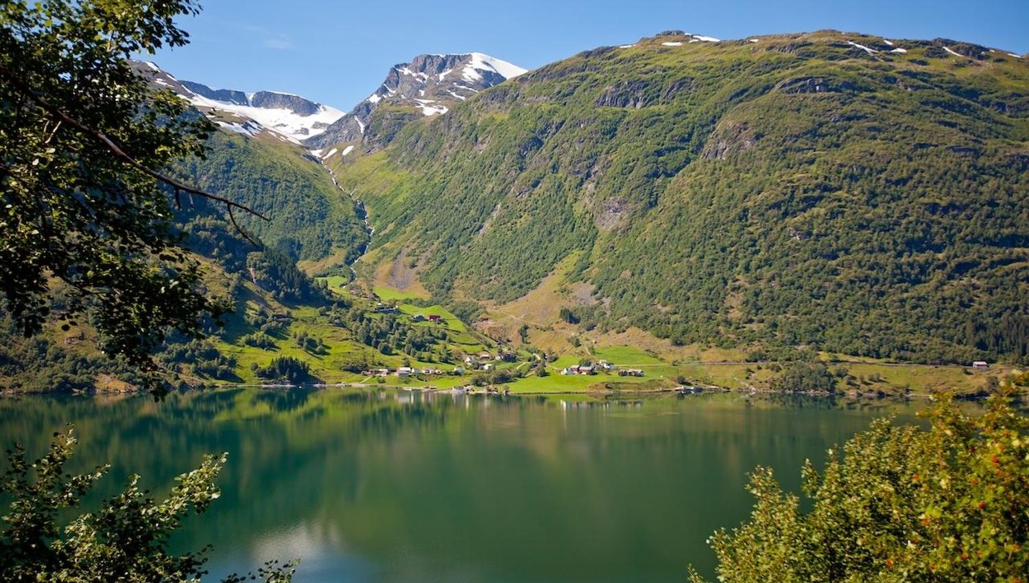 Kandal, Gloppen -Green mountains with snow patches rise above a calm lake, with a small village on the shore under blue sky..