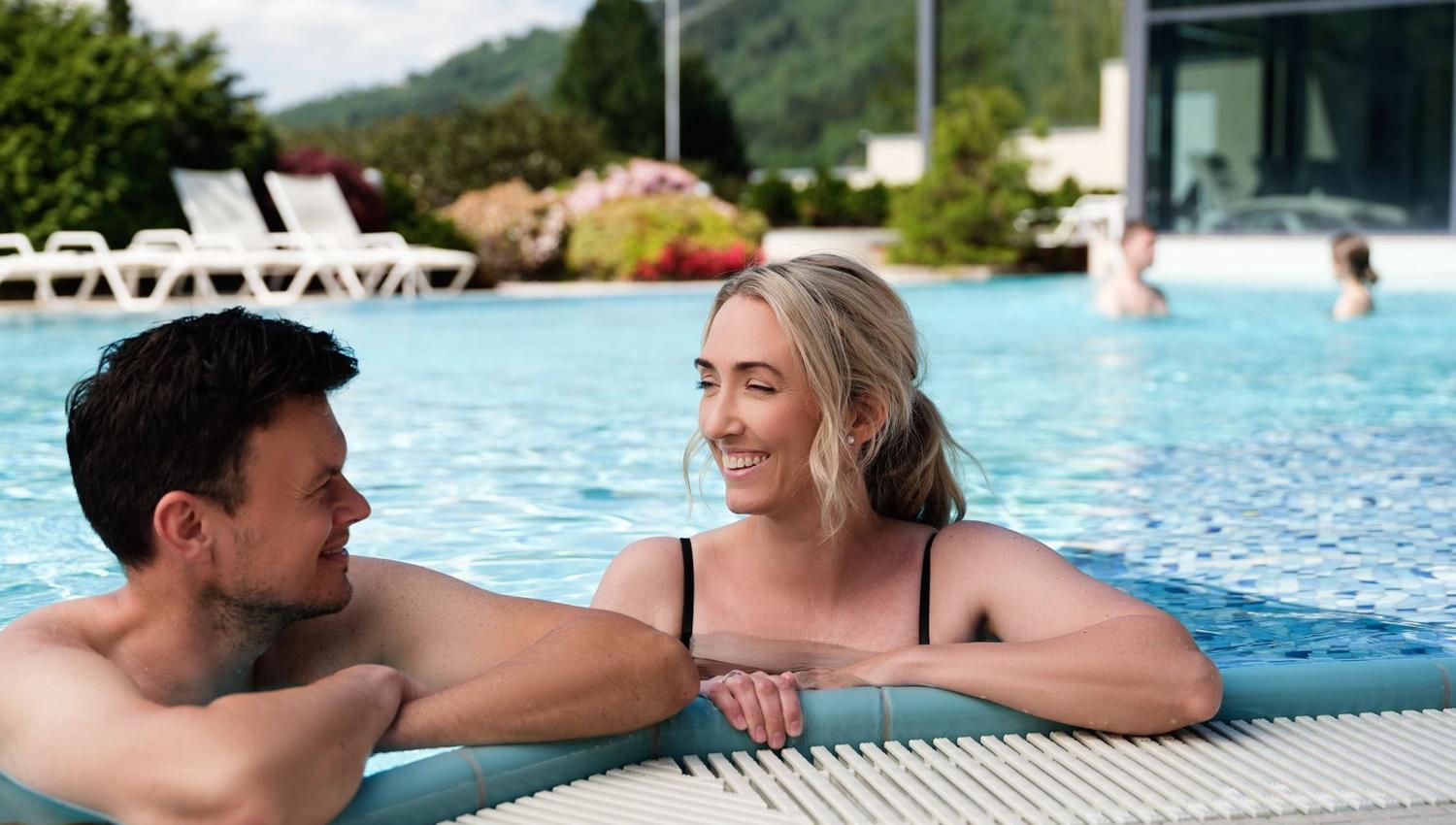 Two people lean on the edge of a blue outdoor pool, with lounge chairs, greenery, and mountains behind..