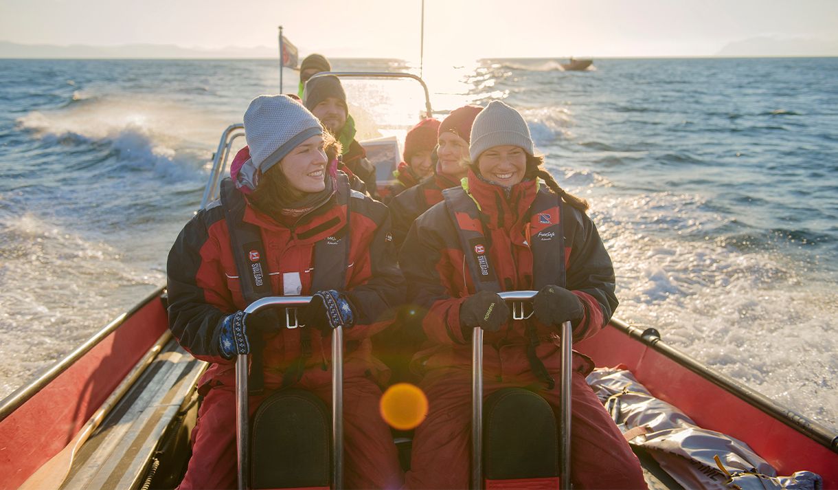 En gruppe med gjester om bord en RIB båt som kjører i fart på en fjord med solskinn over fjorden i bakgrunnen