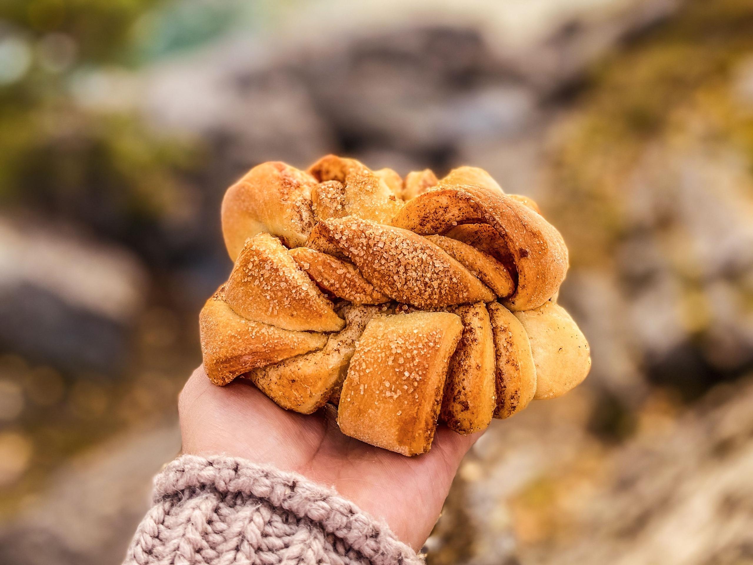 Close-up of a hand in a knitted sleeve holding a twisted sugar-topped pastry against a blurred outdoor background..