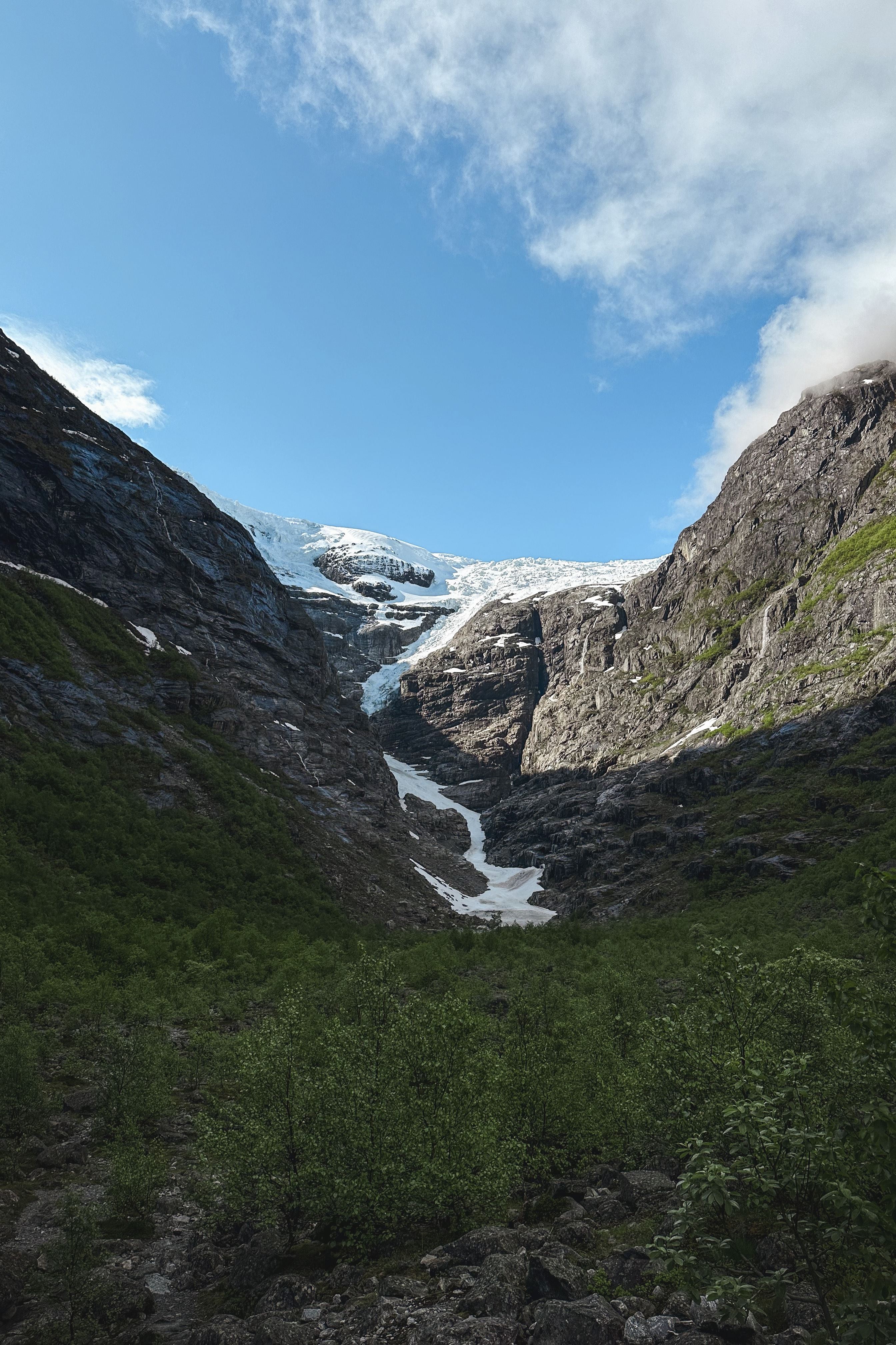 Rocky mountain valley with green shrubs leading to a distant glacier and snow patches beneath a blue sky..