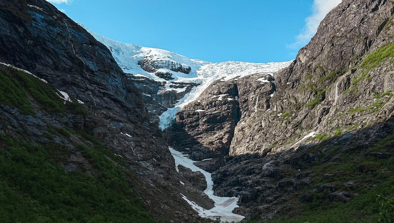 Rocky mountain valley with green shrubs leading to a distant glacier and snow patches beneath a blue sky..