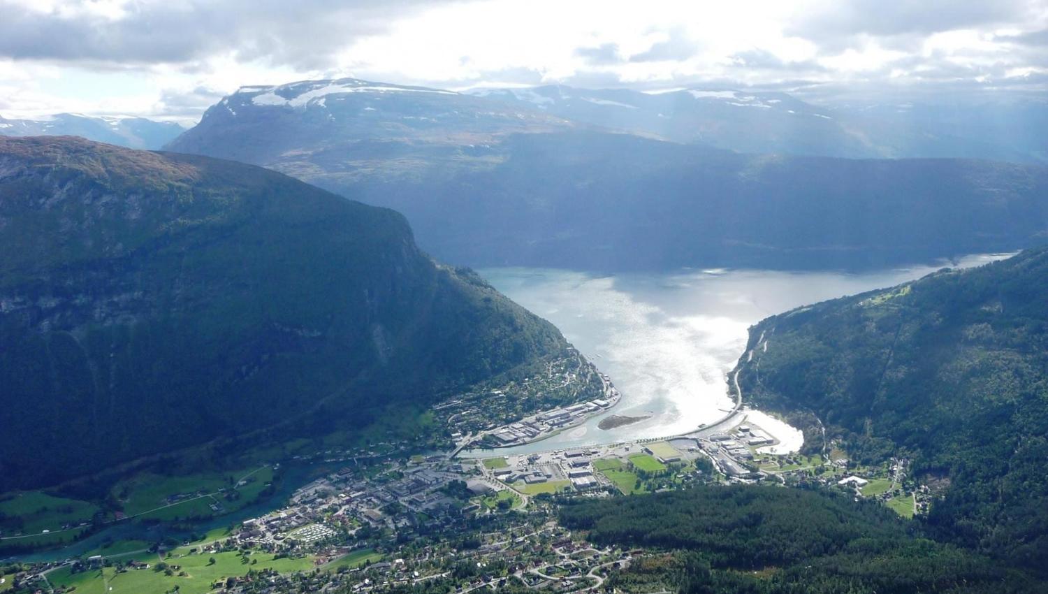 Aerial view of a town and green valley beside a winding lake, surrounded by forested mountains under cloudy sky.