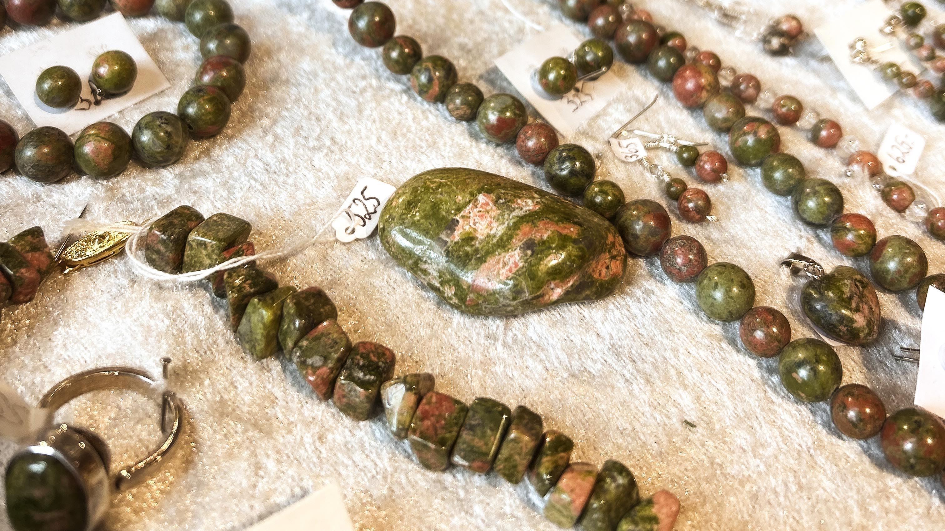 Assorted polished green and pink stone bead jewelry and a large oval stone arranged on a white fabric surface..