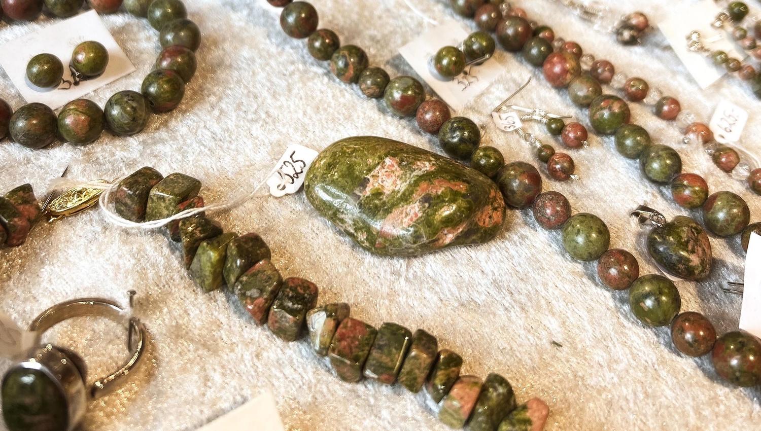 Assorted polished green and pink stone bead jewelry and a large oval stone arranged on a white fabric surface..