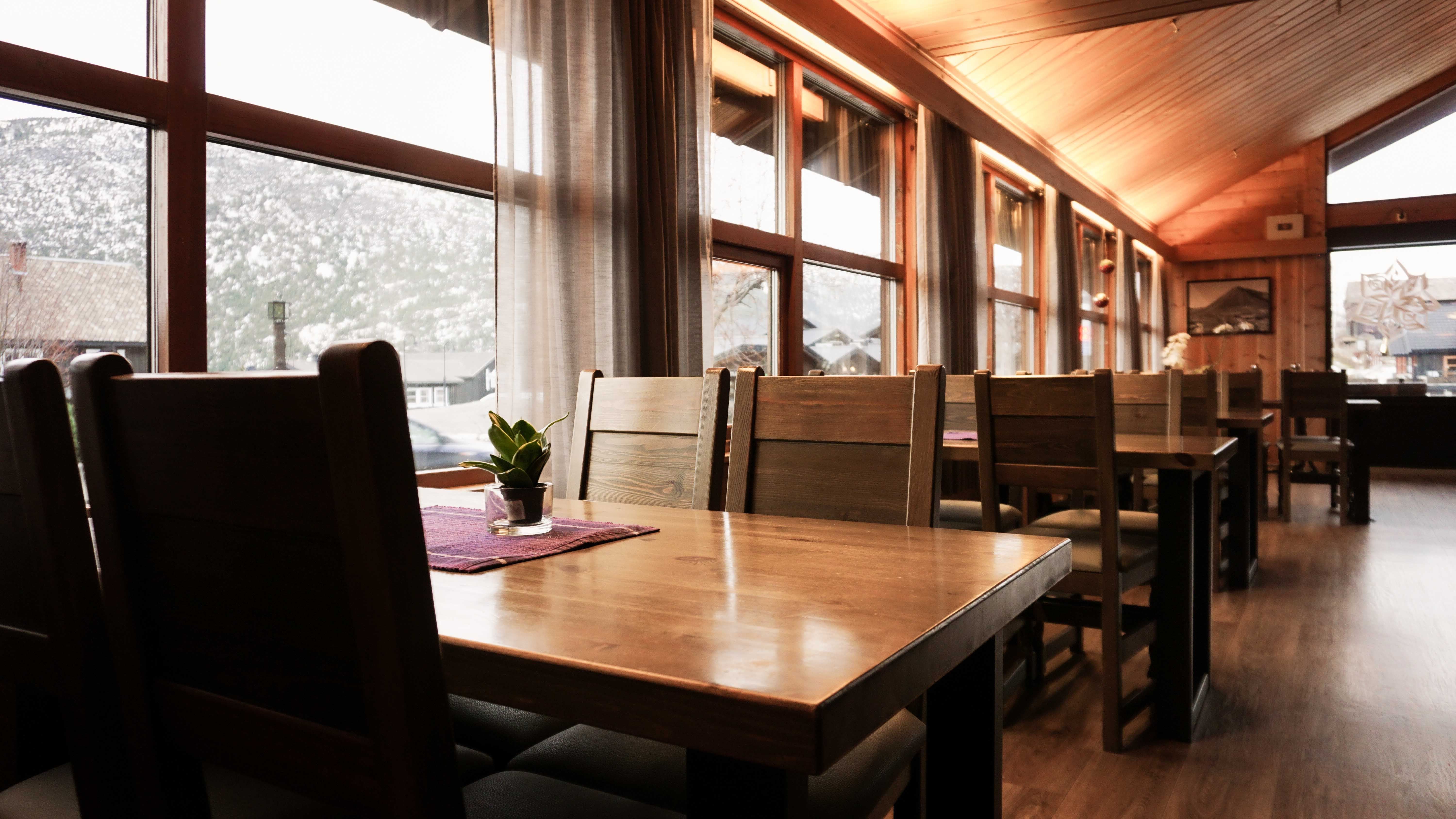 Warm wooden dining area with empty tables and chairs beside tall windows, with mountains and buildings outside..