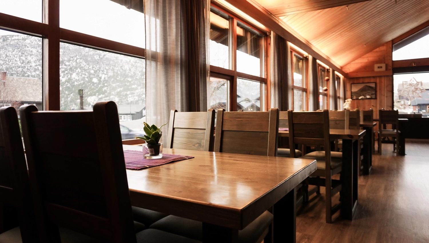 Warm wooden dining area with empty tables and chairs beside tall windows, with mountains and buildings outside..