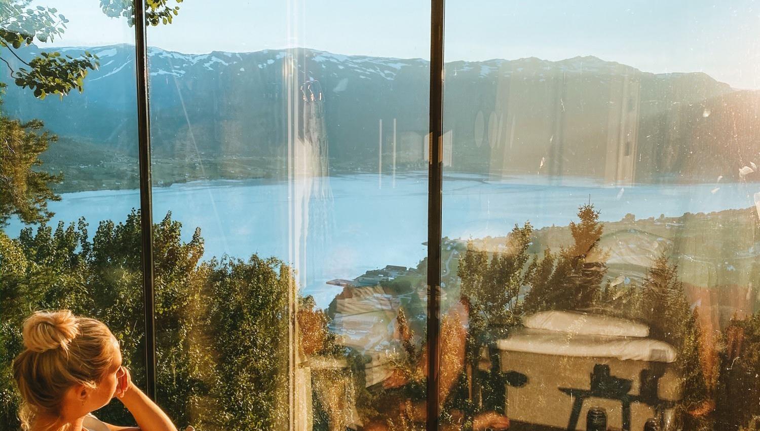 Person lounging in a chair by a large window, overlooking a sunlit lake, forested hills, and mountains..