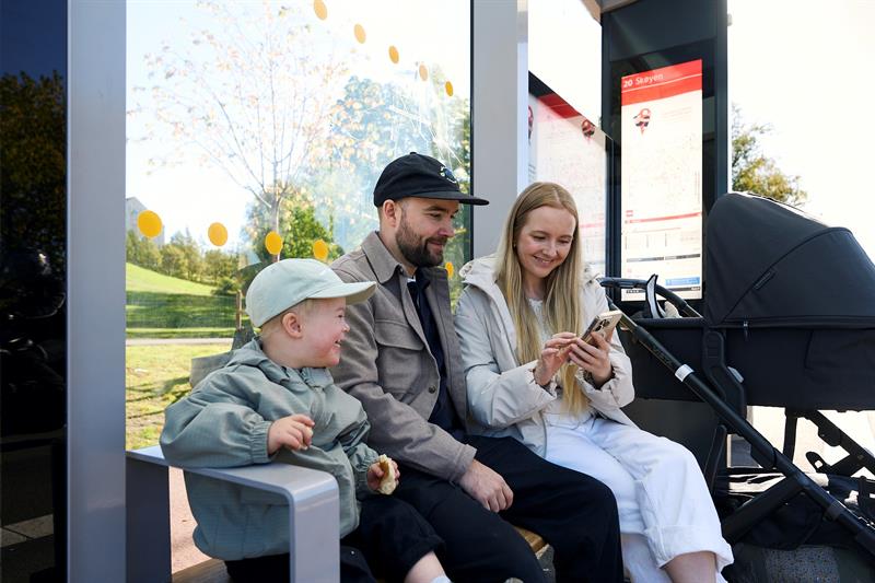 Mother, father and a child sitting on a bench at a bus stop with a stroller, looking at a mobile phone.