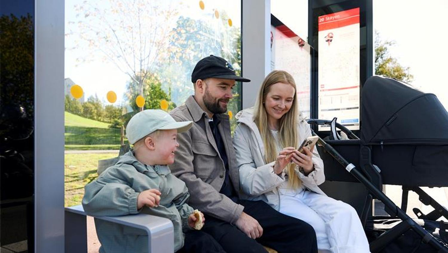 Mother, father and a child sitting on a bench at a bus stop with a stroller, looking at a mobile phone.