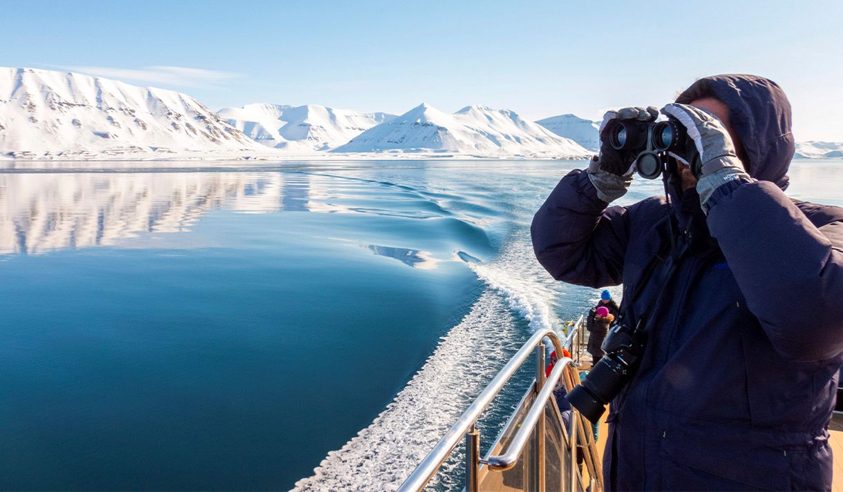 A guest on a boat in the foreground scouting out across a calm blue fjord and mountains with binoculars
