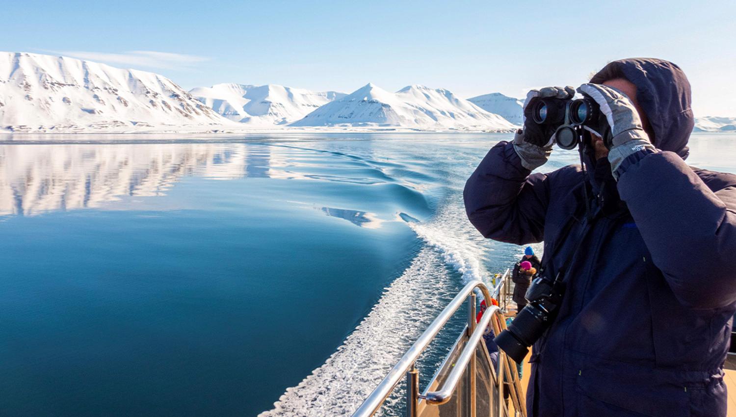 A guest on a boat in the foreground scouting out across a calm blue fjord and mountains with binoculars