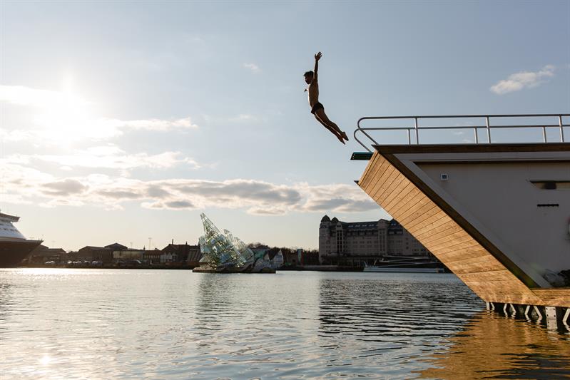 a person plunging from the roof of one of the saunas into the fjord, Oslo's panorama in the background