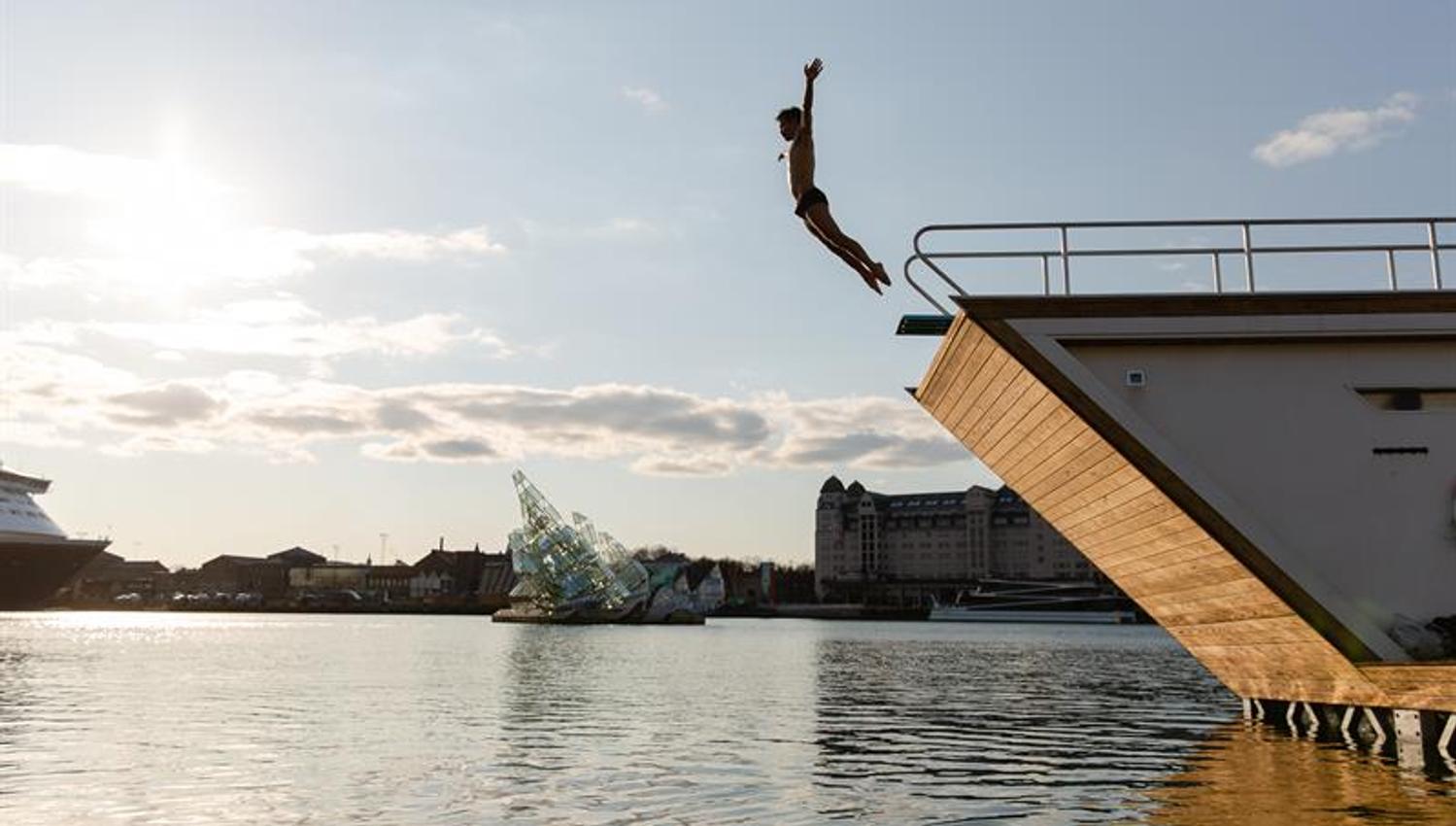 a person plunging from the roof of one of the saunas into the fjord, Oslo's panorama in the background