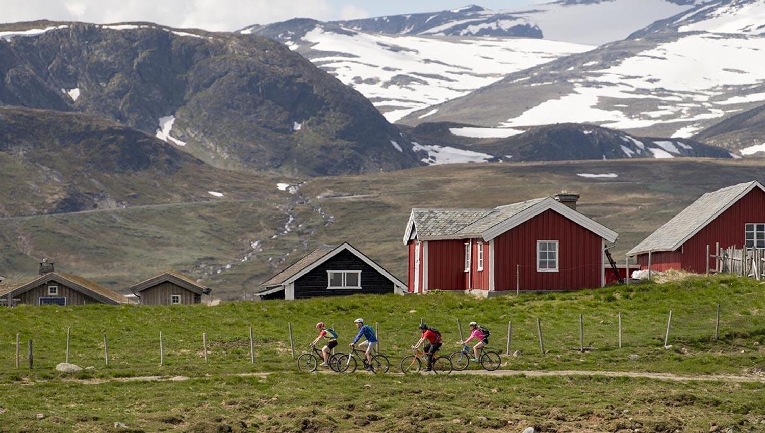 Cyclists in fron of red farm houses with a dramatic mountain scenery in the background.