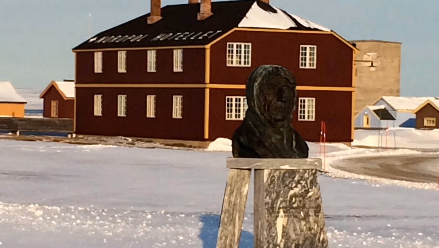 A metal bust of Roald Amundsen in the centre of Ny-Ålesund, with buildings and snowy streets in the background