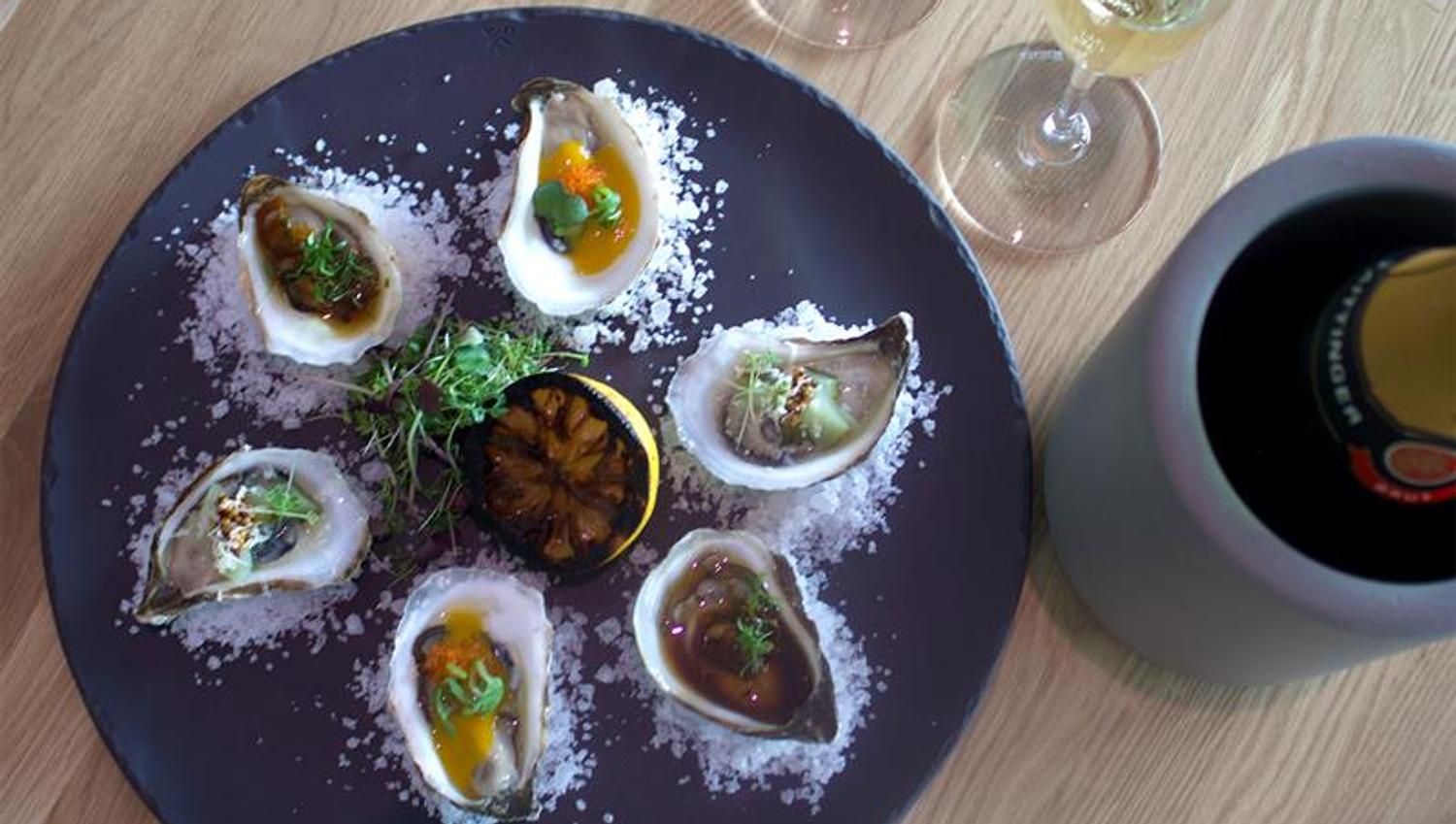 Detail of oyster on a plate with salt sprinkled around and half a lemon in the middle. Two glasses of sparkling white wine next to it.
