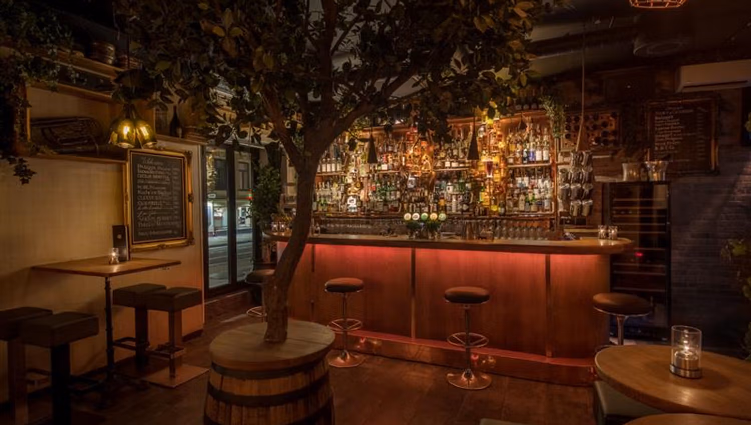 Bar interior with bar counter, bar stools, small tables and chairs, and a tree standing in a barrel in the middle of the room.