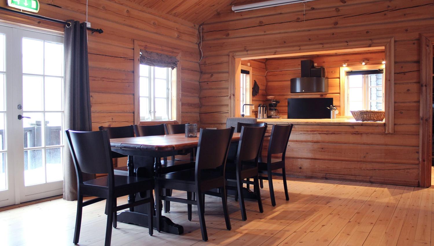 Interiör i timmerstuga med lång matbord, svarta stolar, fönster och serveringslucka mot ett kök.