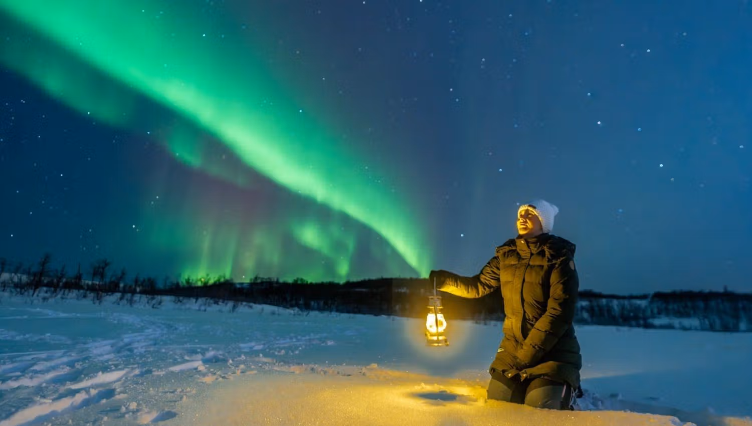 A person holding a lantern in a snowy landscape beneath the Northern Lights during a Northern Lights chase from Tromsø.