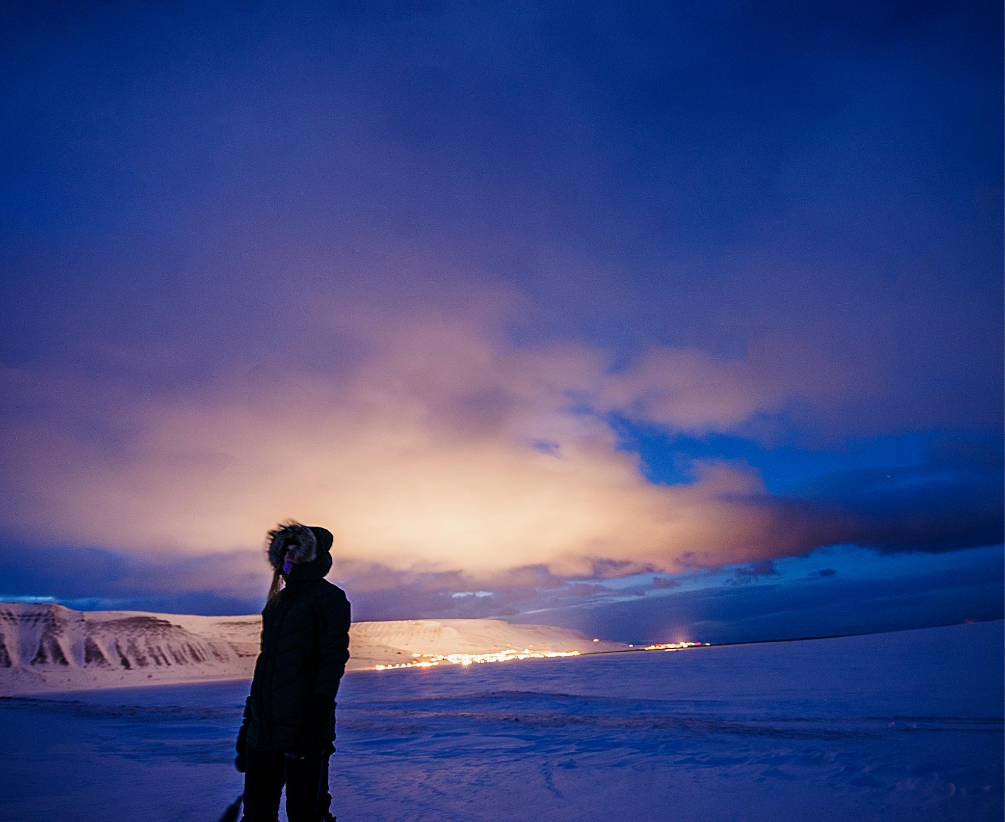Person standing in front, on a dark winter day with the lights from town in the background 