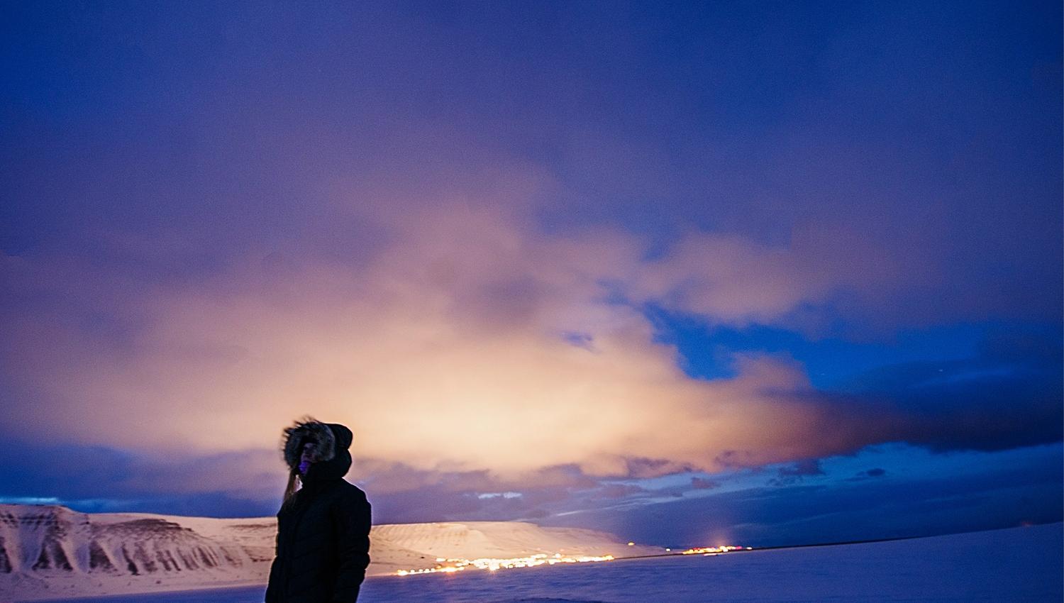 Person standing in front, on a dark winter day with the lights from town in the background