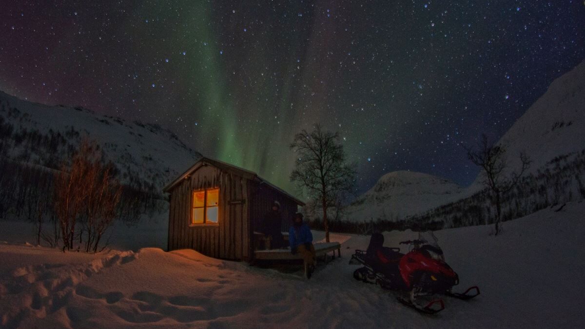 A small cabin and a snowmobile under the northern lights