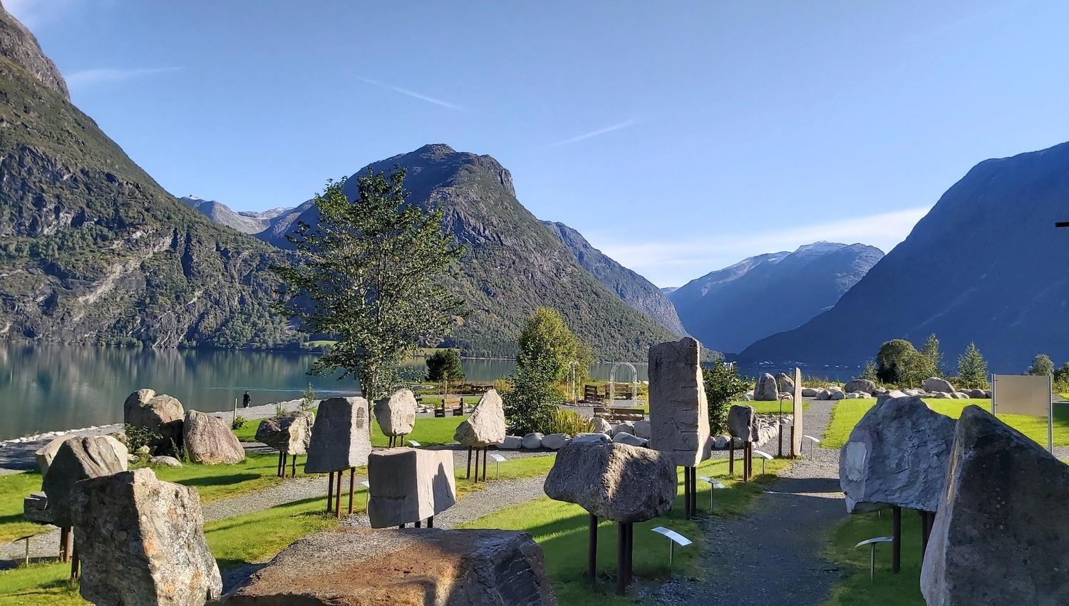 Store steinblokker på stativ i ein park ved ei stille fjord, med bratte fjell og blå himmel i bakgrunnen..