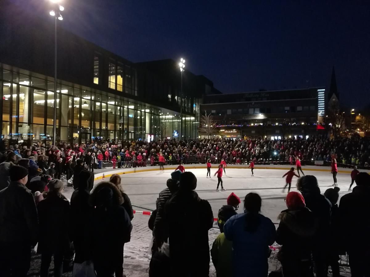 An outdoor ice rink in the city center at night, surrounded by spectators and illuminated by bright floodlights next to Hamar cultural center