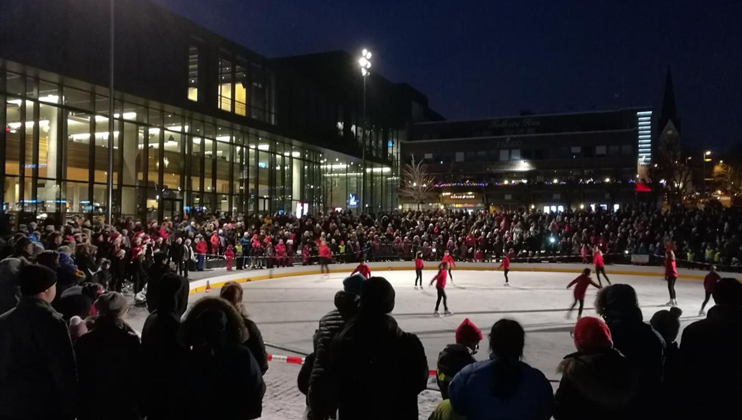An outdoor ice rink in the city center at night, surrounded by spectators and illuminated by bright floodlights next to Hamar cultural center
