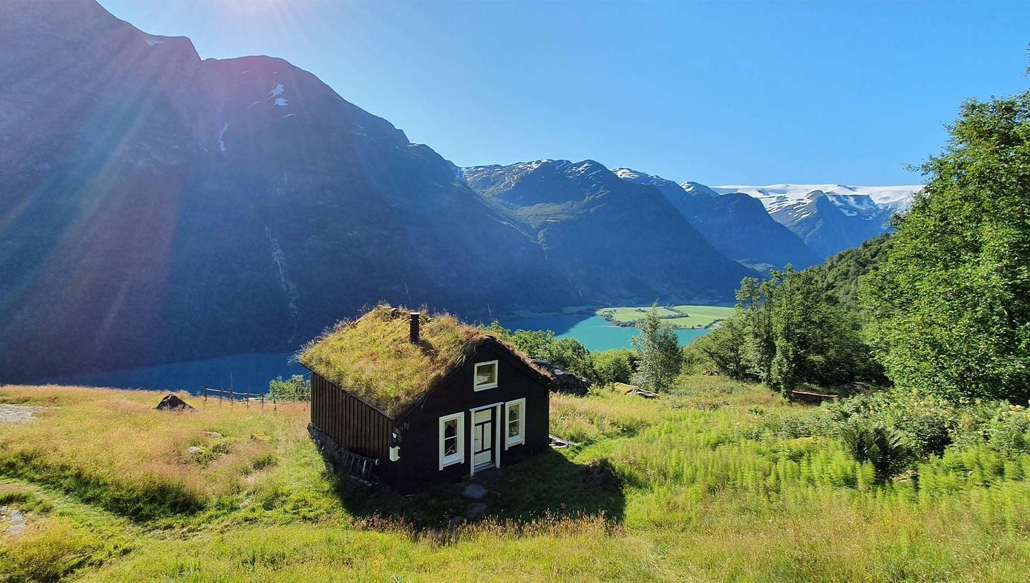 Small dark cabin with a grass roof on a sunny hillside, overlooking a blue fjord and snow-capped mountains.