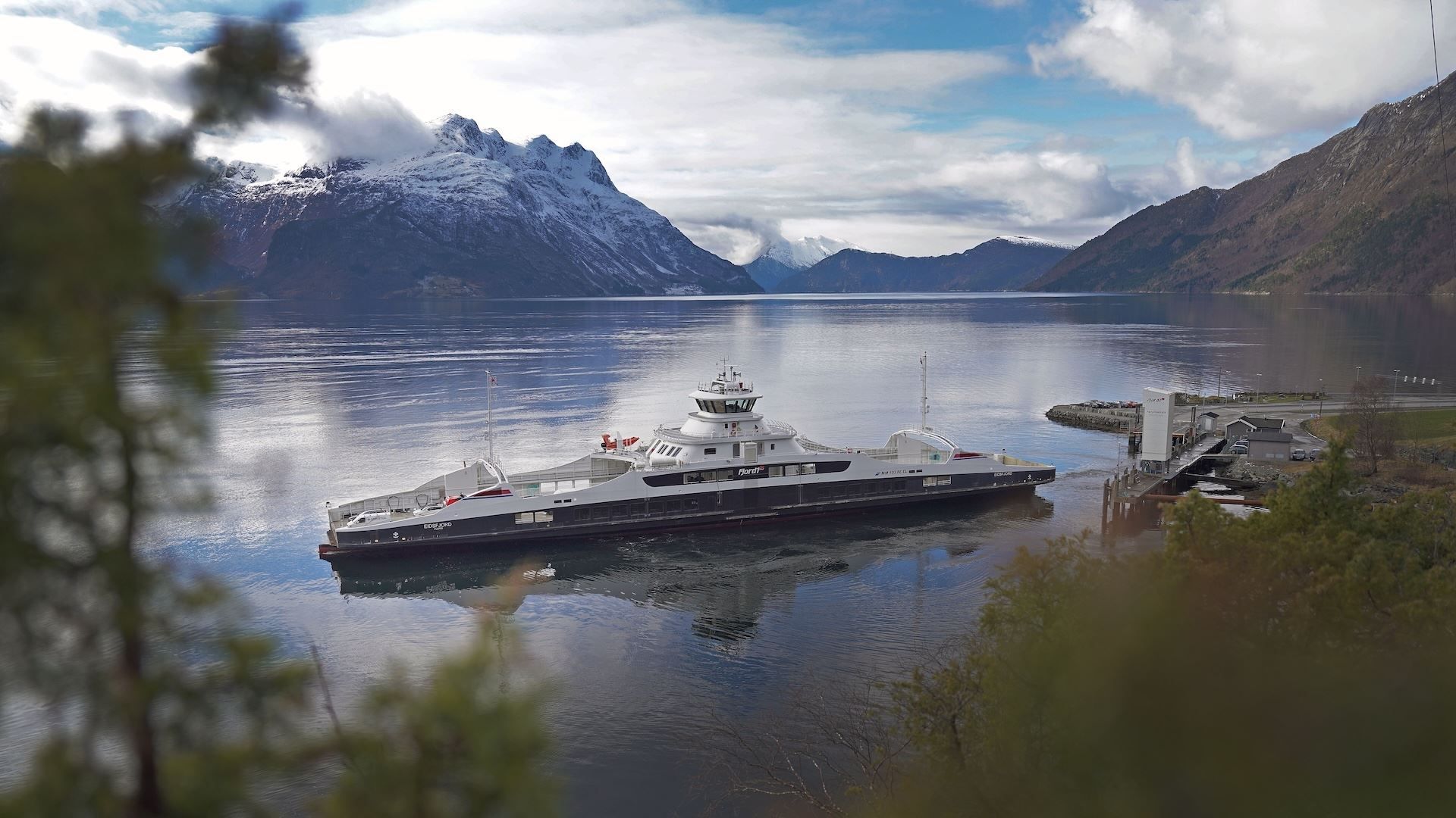 A large Fjord1 ferry docked at a small pier on calm water, with snow-capped mountains and clouds in the distance..