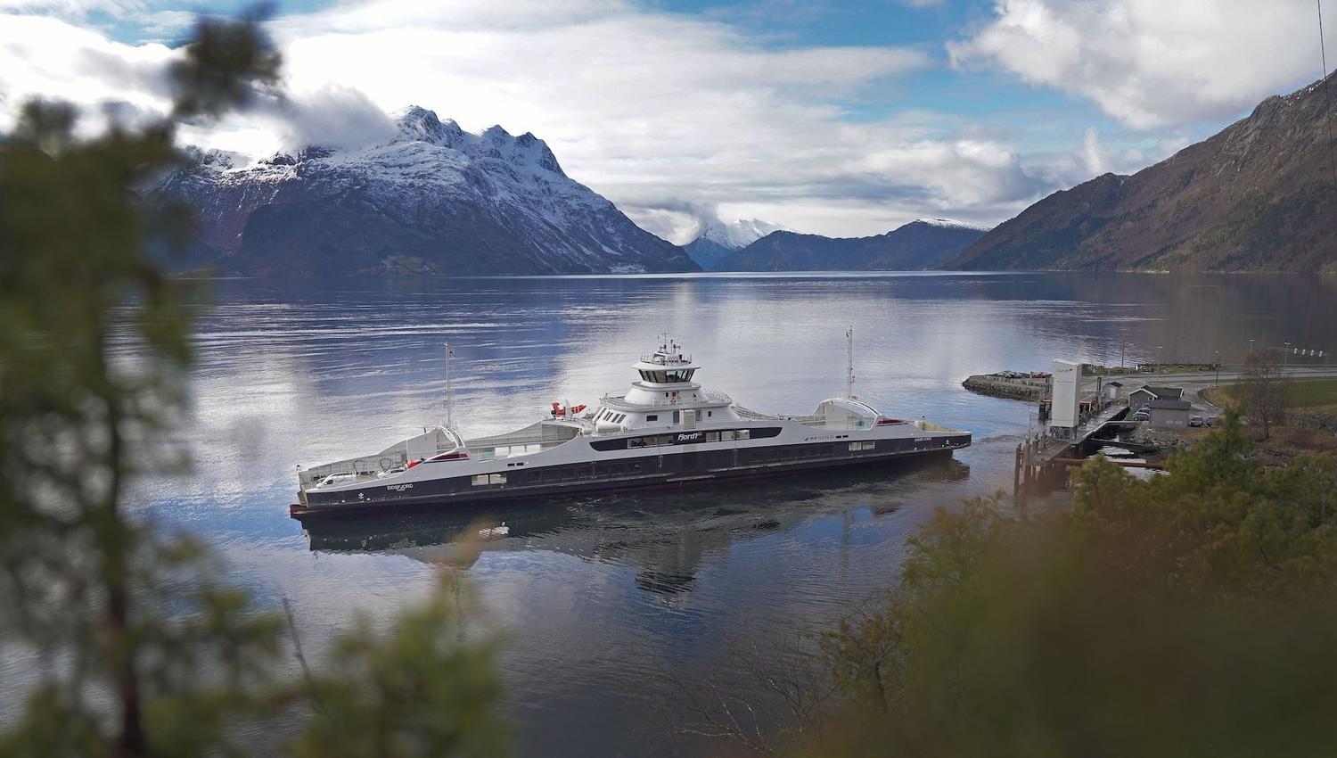 A large Fjord1 ferry docked at a small pier on calm water, with snow-capped mountains and clouds in the distance..