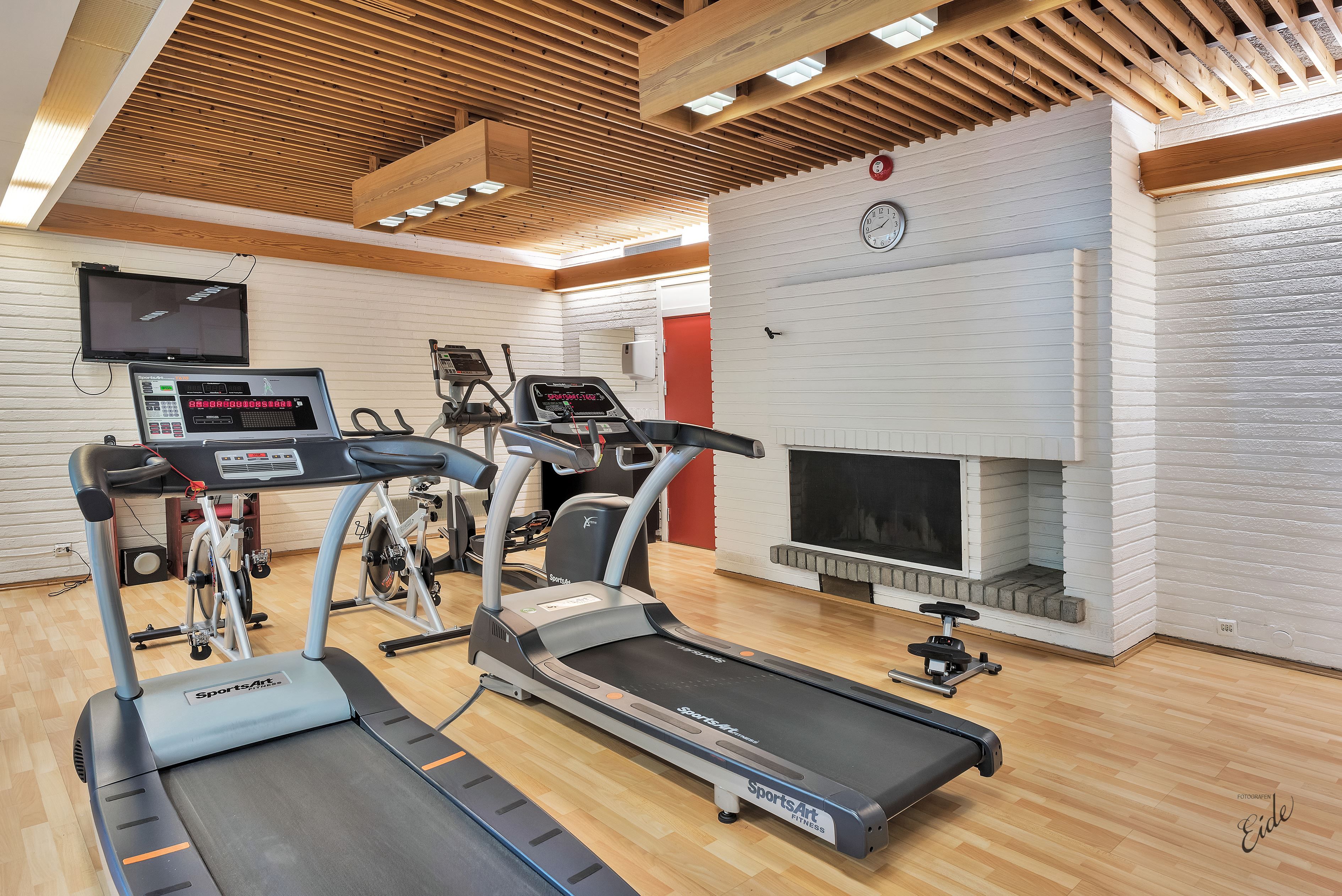 Bright fitness room with two treadmills, a stationary bike, wooden slat ceiling, TV, and a white brick fireplace.