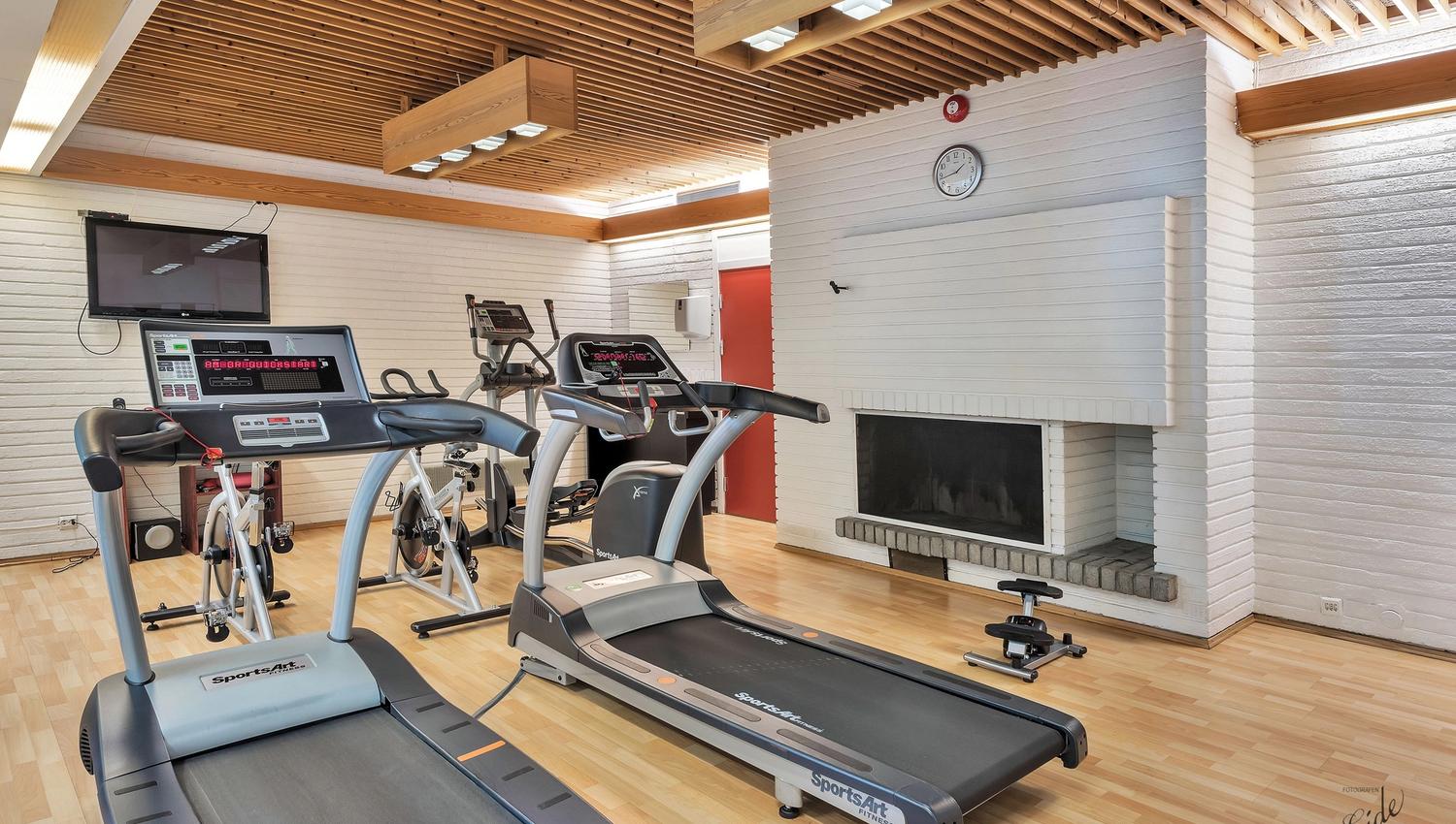 Bright fitness room with two treadmills, a stationary bike, wooden slat ceiling, TV, and a white brick fireplace.