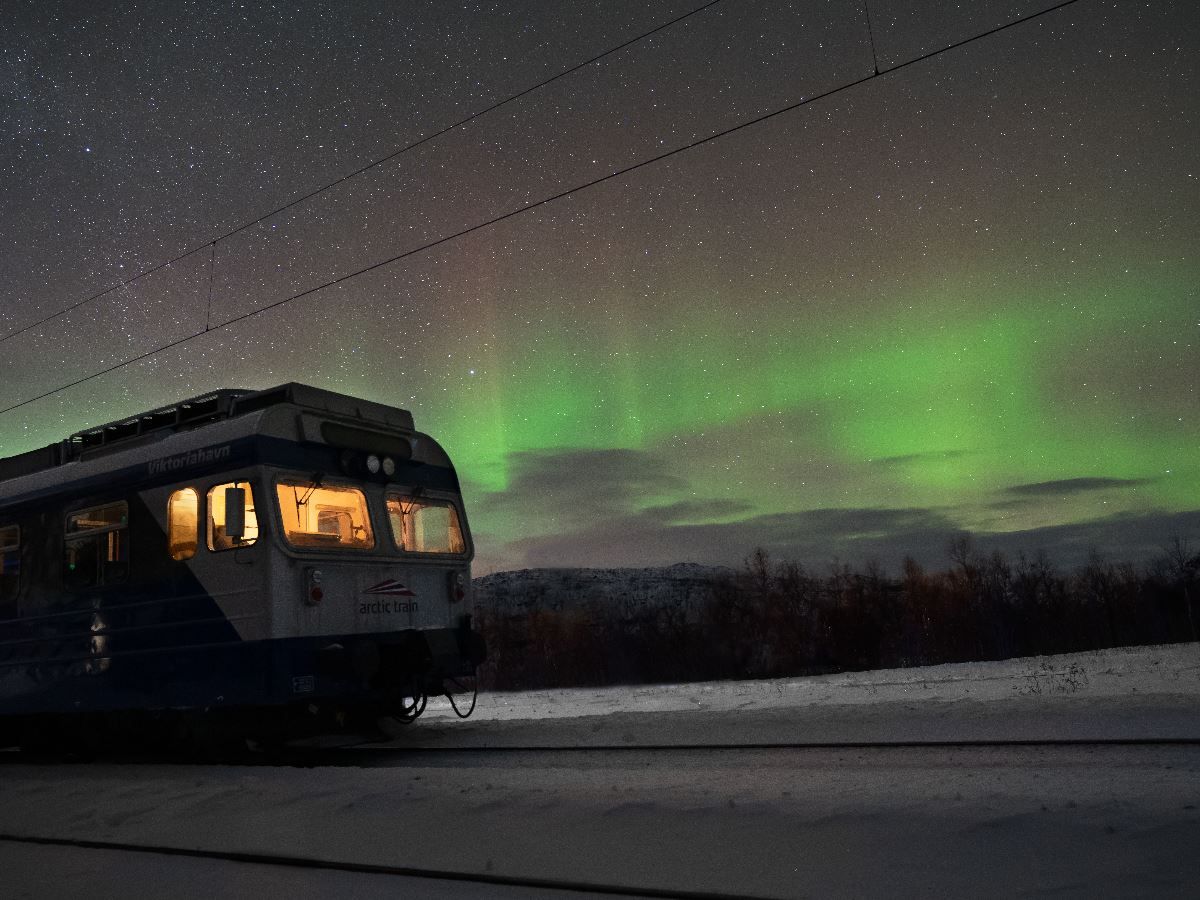 A passenger train on snowy tracks at night with glowing cabin lights beneath a starry sky and green aurora..