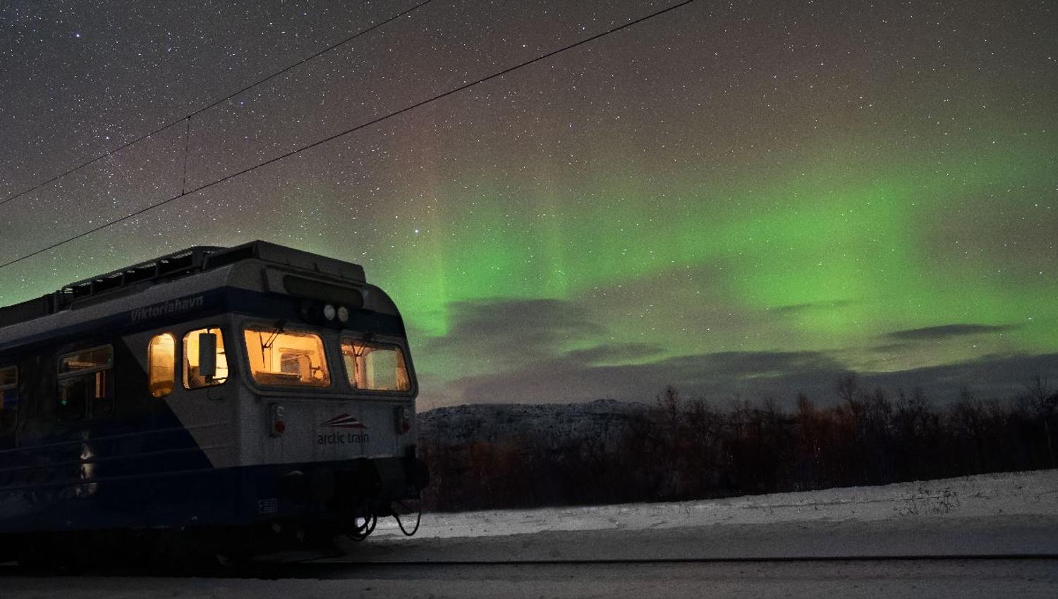 A passenger train on snowy tracks at night with glowing cabin lights beneath a starry sky and green aurora..