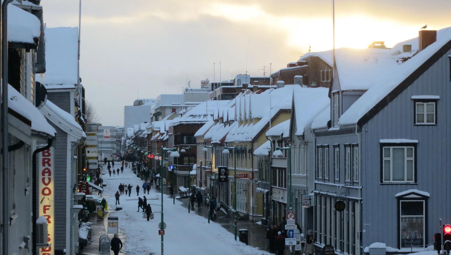 Snow-covered town street with people, cars, and buildings under a cloudy sky with a glimpse of sunlight.