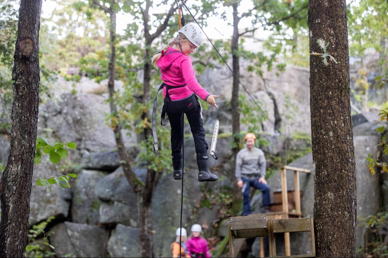A young girl hangs from a zip line between the trees while an adult stands ready to receive her