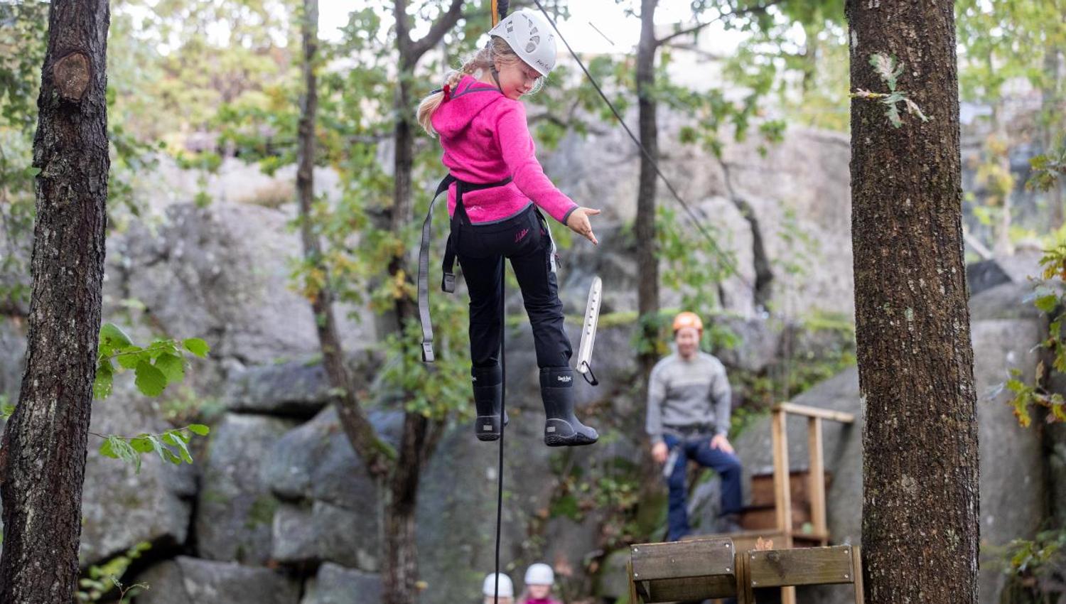 A young girl hangs from a zip line between the trees while an adult stands ready to receive her
