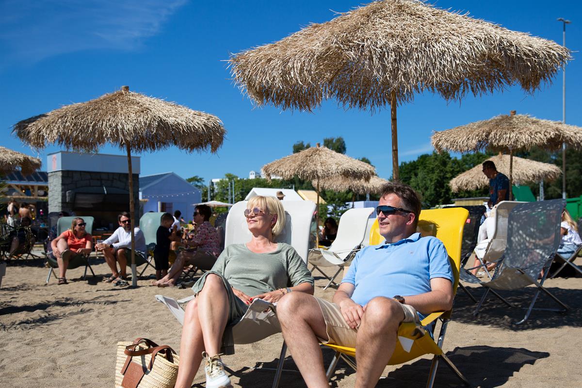 Personer som slapper av i solstoler under store stråparasoller på stranden.
