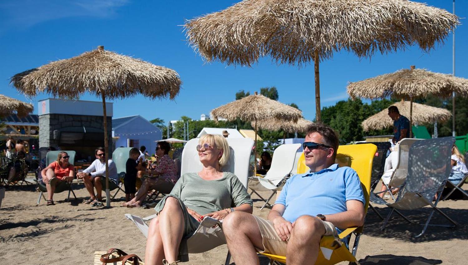 Personer som slapper av i solstoler under store stråparasoller på stranden.