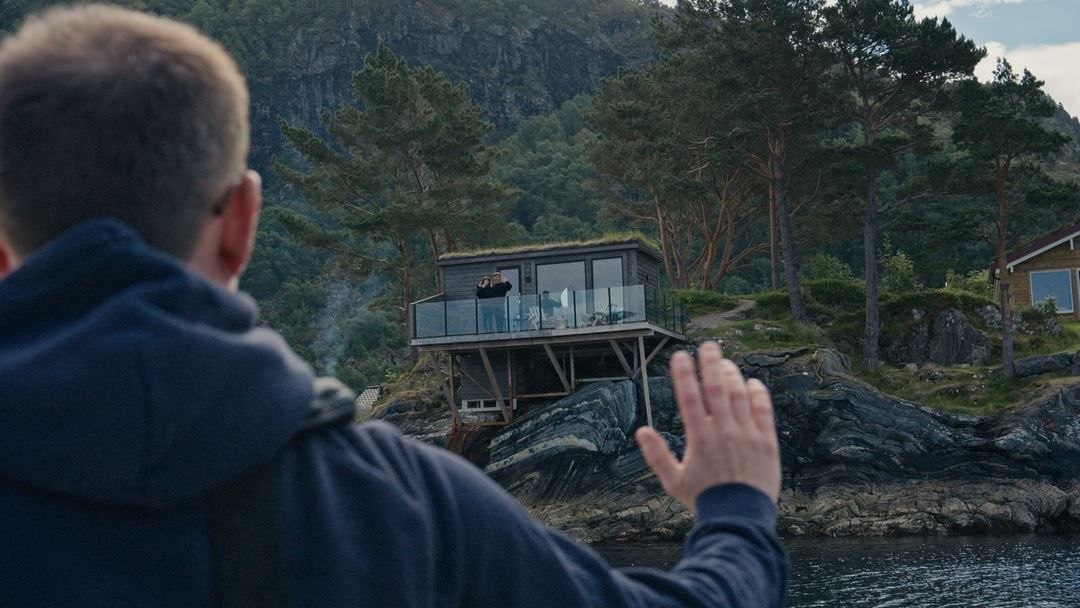 Over-the-shoulder view of a person waving toward a modern cabin on a rocky lakeshore amid pine trees and hills..