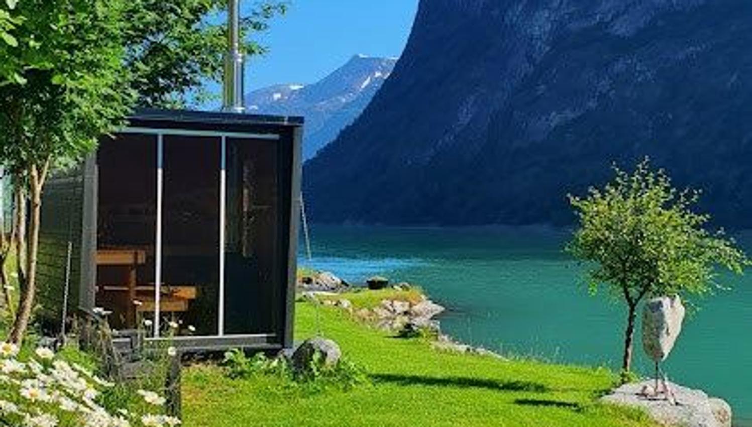 Small cabin on a grassy lakeshore with white daisies, turquoise water, and a steep mountain under clear blue sky.
