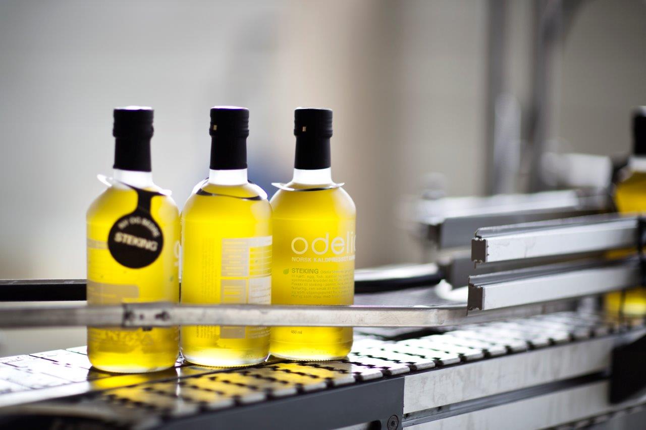 Bottles of golden-yellow apple juice stand on a conveyor belt in a production facility. The labels read “Odel” and “Askim Frukt og bærpresseri.