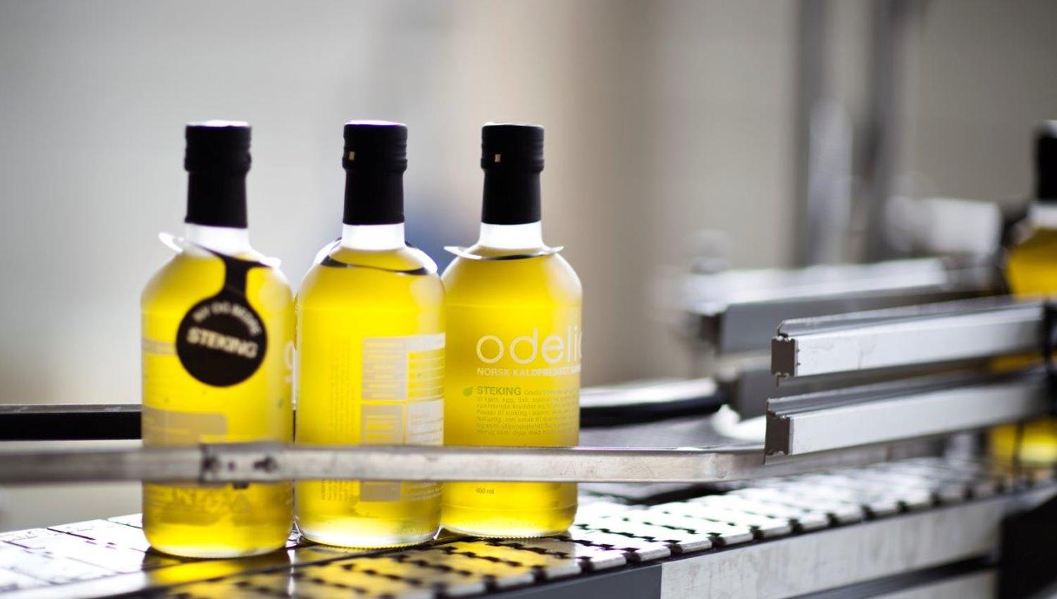 Bottles of golden-yellow apple juice stand on a conveyor belt in a production facility. The labels read “Odel” and “Askim Frukt og bærpresseri.