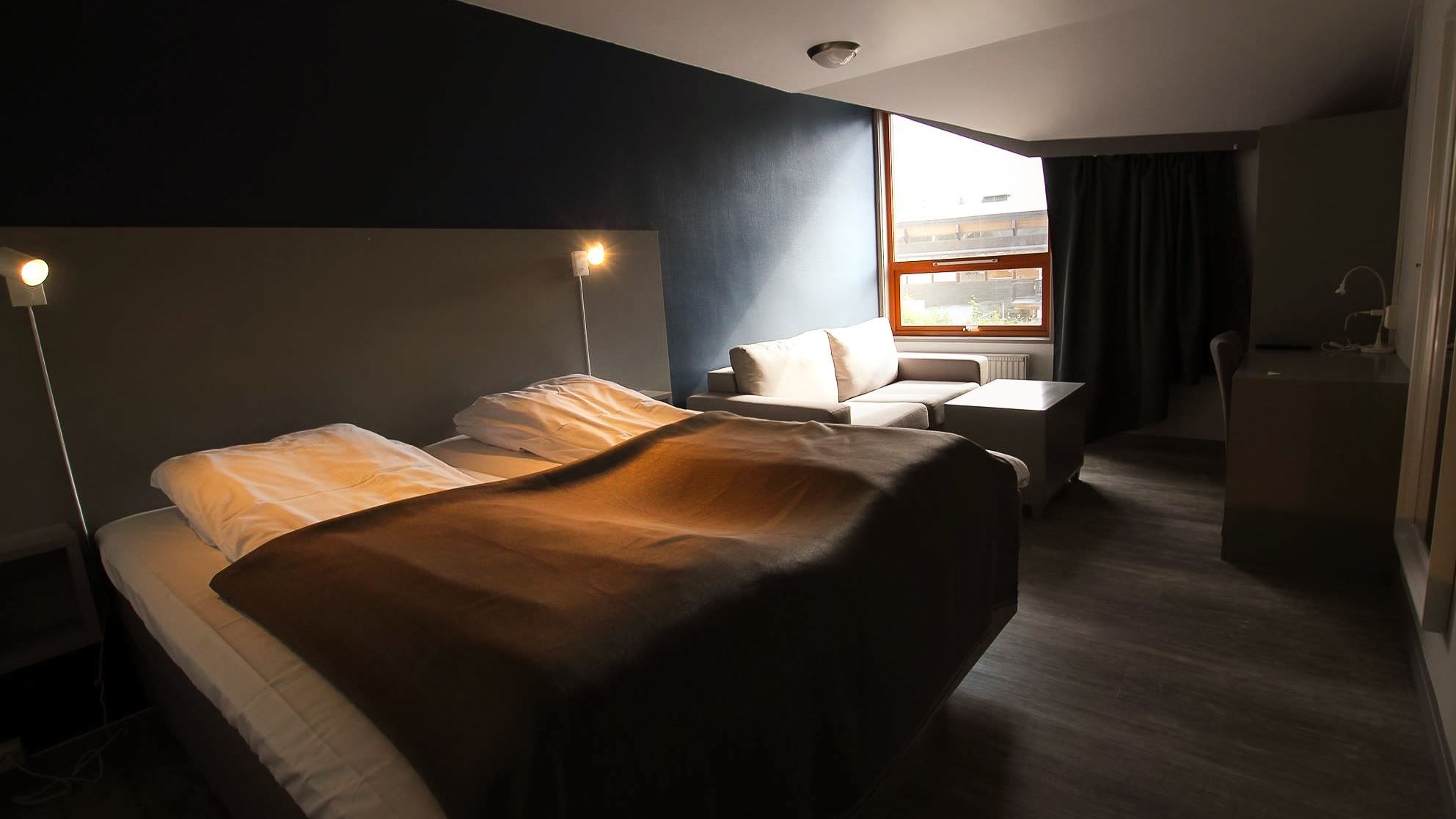 Dimly lit hotel room with an unmade bed, wall lamps, a small sofa, desk, and a window with curtains..