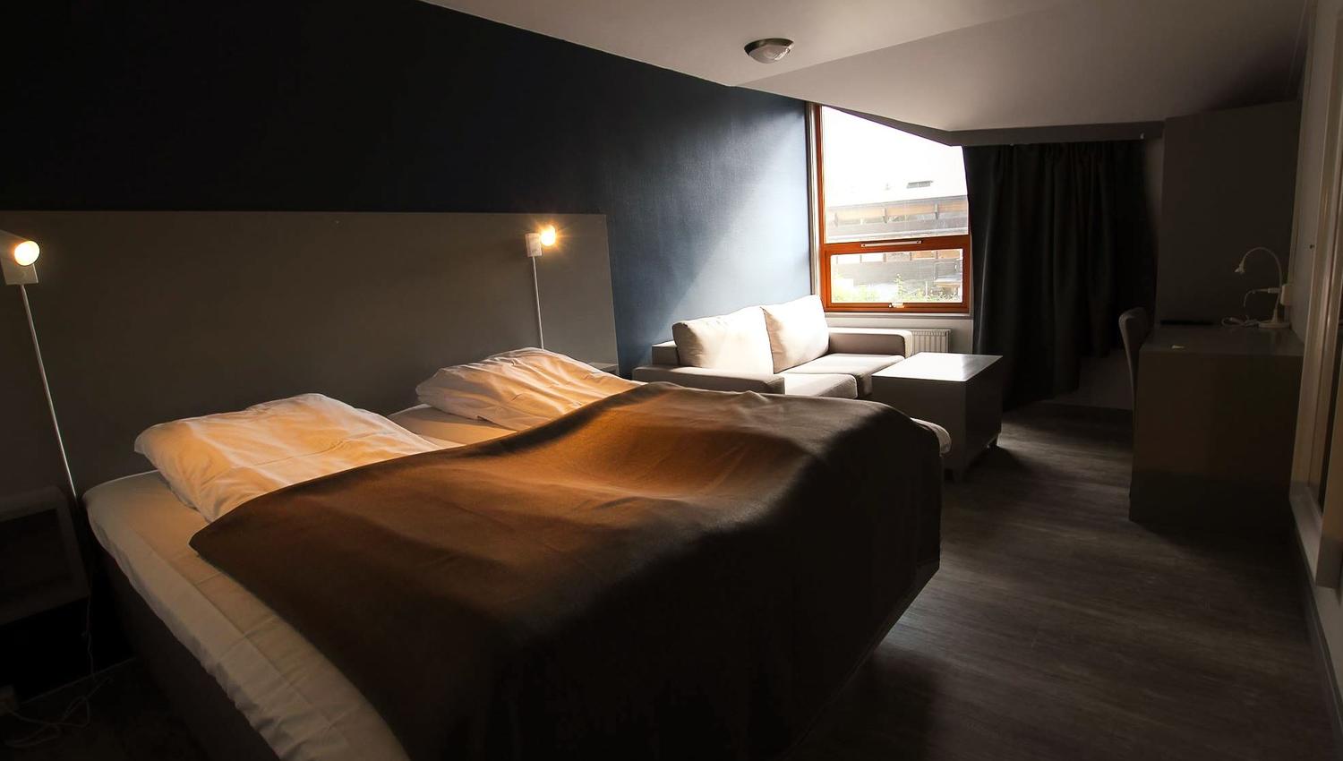 Dimly lit hotel room with an unmade bed, wall lamps, a small sofa, desk, and a window with curtains..